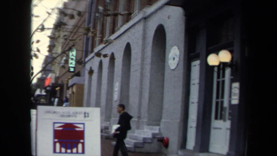 1970 Washington, DC street frame with a man walking past the arched theatre frontage, signs, steps, and globe lights.
