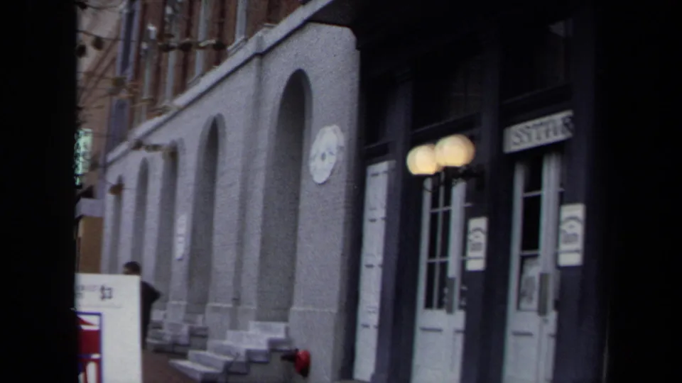 1970 Washington, DC home-movie frame showing arched building entrances, white doors, globe lamps, and a partial sign above one doorway.