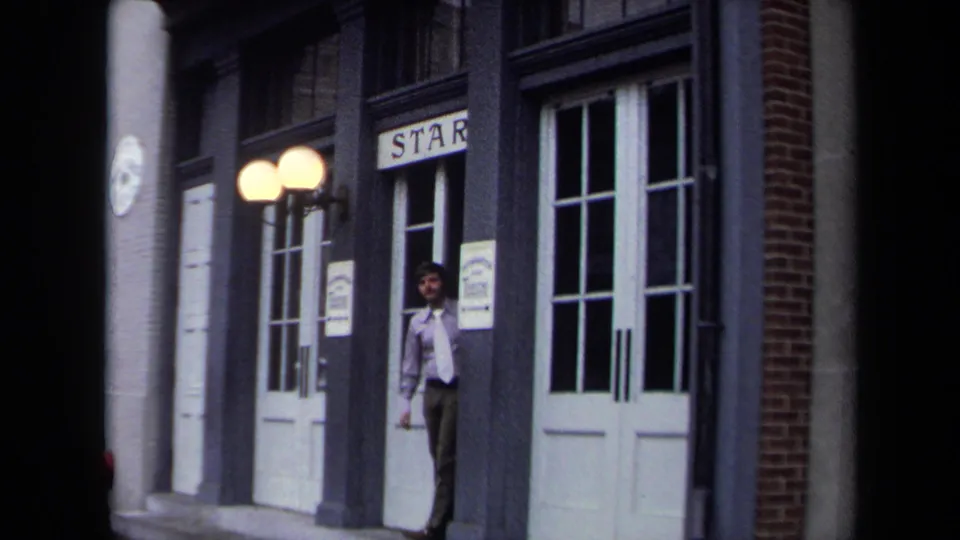 1970 Washington, DC frame showing a man in a white shirt at a doorway near white double doors, globe lamps, and the visible "STAR" sign.