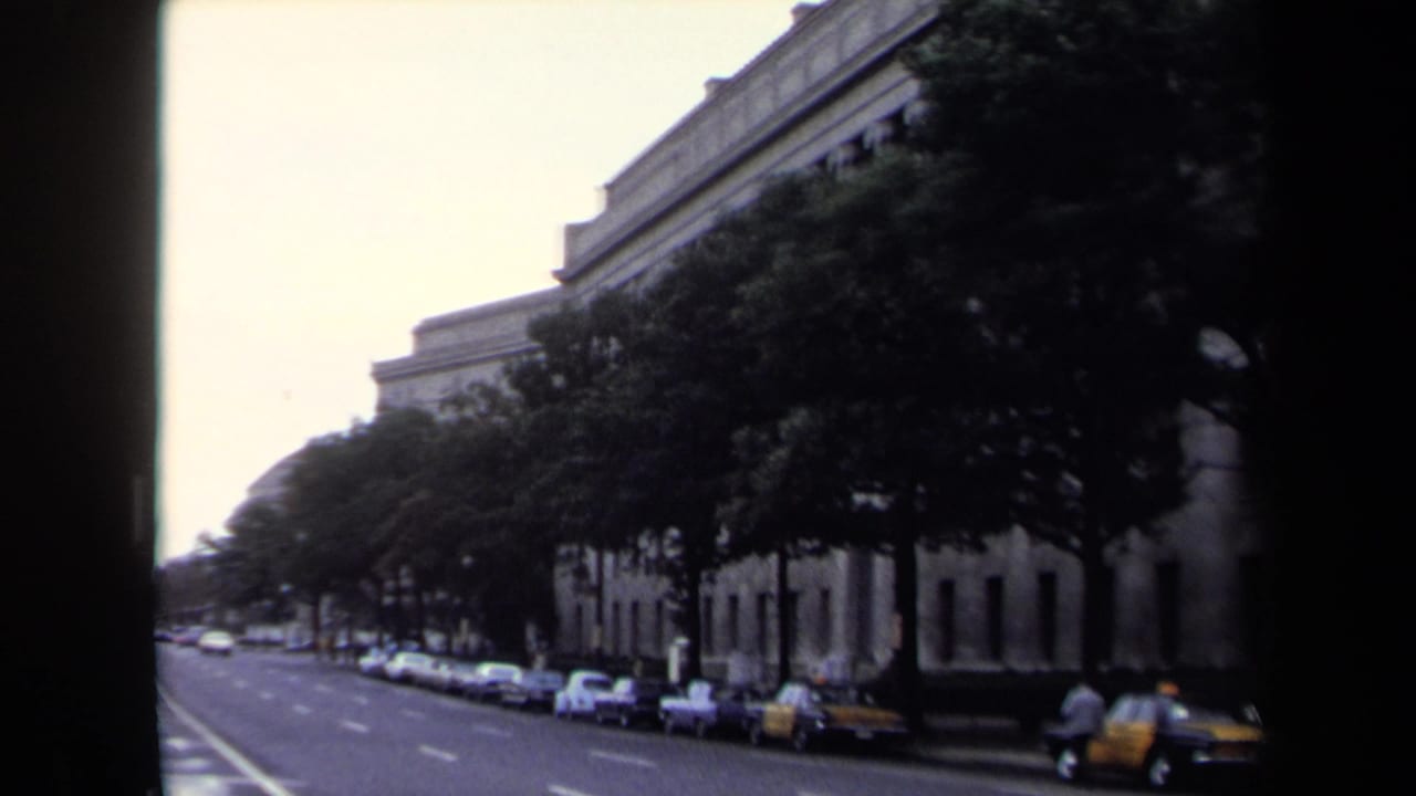 1970 home-movie frame of a wide Washington, DC street bordered by trees, parked cars, and a large building facade.