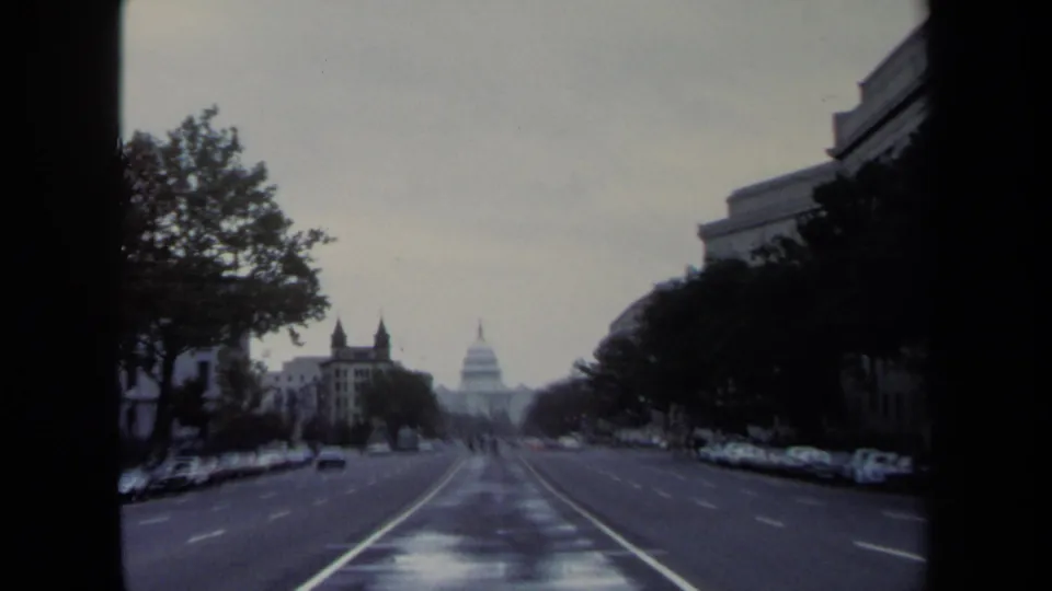 1970 Washington, DC street view with wet pavement, distant Capitol dome, trees, and parked cars along the curb.