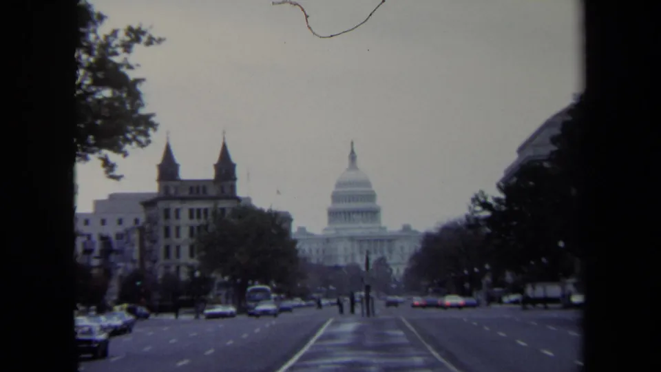 Washington, DC frame facing the U.S. Capitol, with small pedestrians, cars, trees, and older buildings along the avenue.