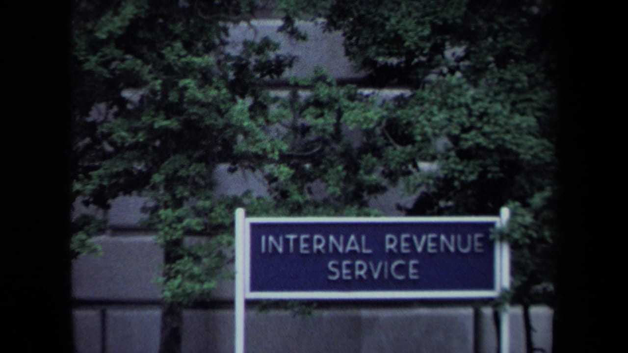 Internal Revenue Service sign in Washington, DC, framed by green trees and the exterior wall of a federal building.