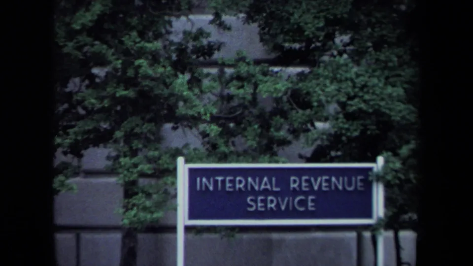 Internal Revenue Service sign in Washington, DC, framed by green trees and the exterior wall of a federal building.