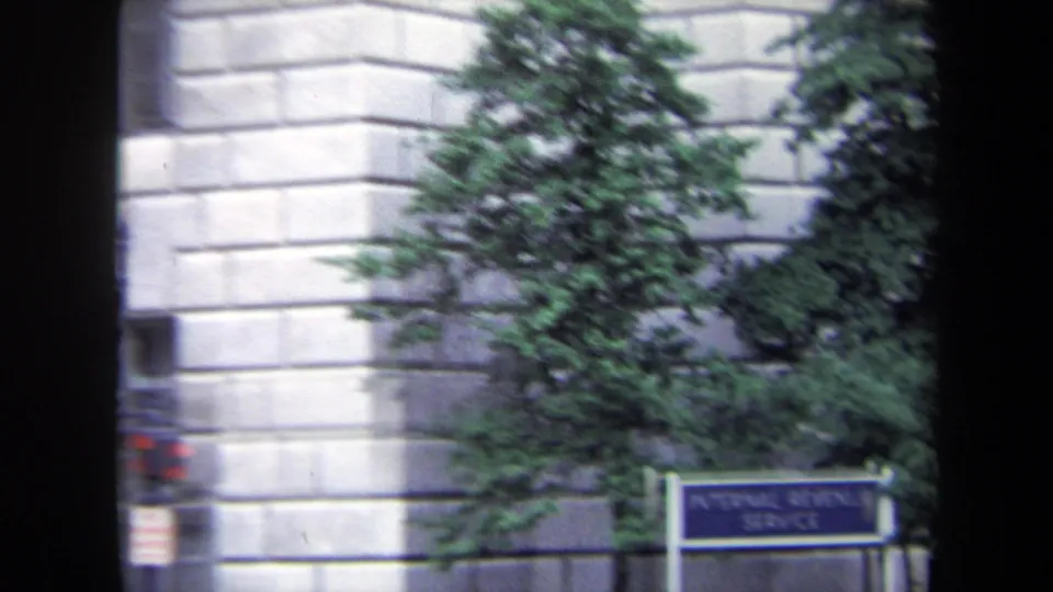A wider, slightly soft view of an Internal Revenue Service sign near a stone facade, trees, and the edge of a city street.