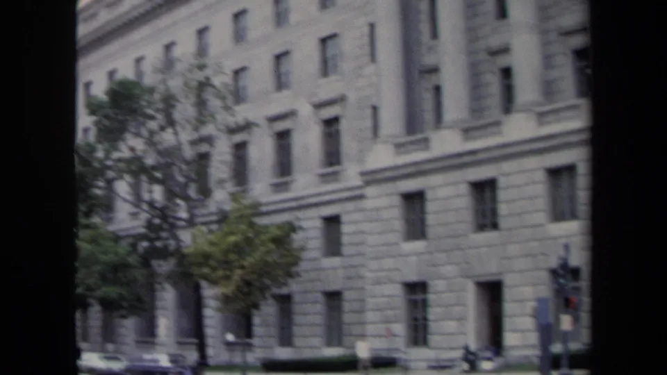 Washington, DC government-building exterior with rows of windows, stonework, trees, and a few street-level details.
