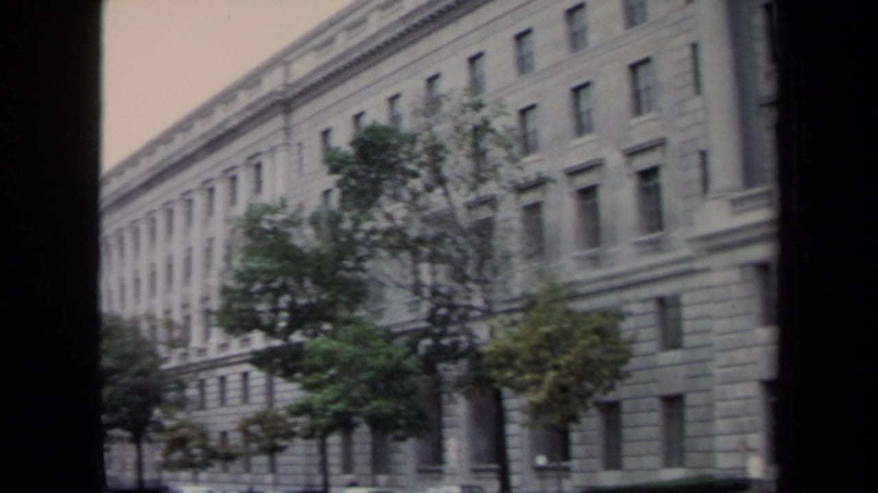 1970 8mm frame of a monumental stone building and street trees beside parked cars in Washington, DC.