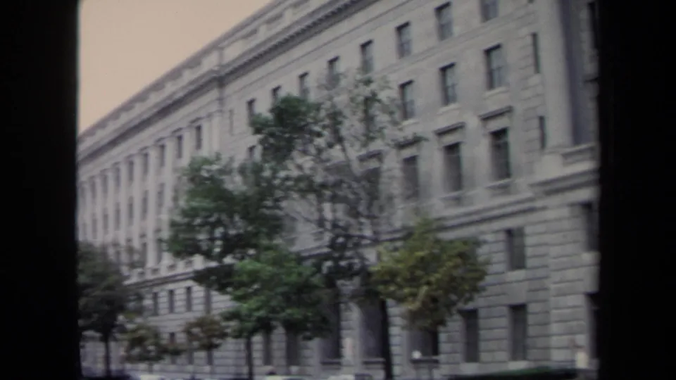 1970 8mm frame of a monumental stone building and street trees beside parked cars in Washington, DC.