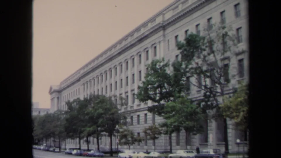 1970 Washington, DC 8mm view of a long monumental facade bordered by trees and parked automobiles.