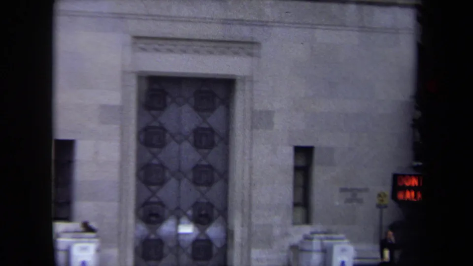 1970 Washington, DC home-movie frame centered on an ornate metal entrance door in a monumental stone facade, with signs and sidewalk details nearby.