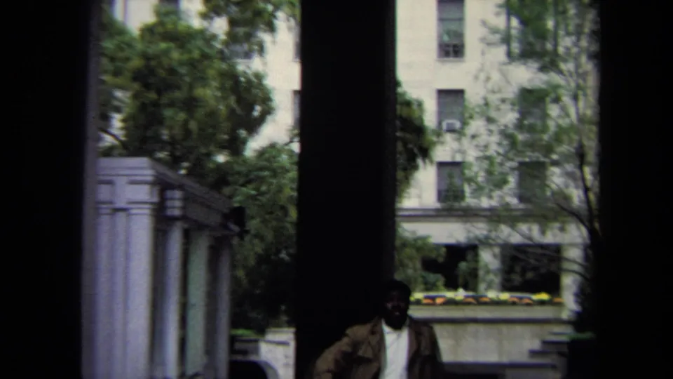 Handheld 8mm frame in Washington, DC showing stone courtyard architecture, trees, and a shadowed pedestrian near the lower edge.
