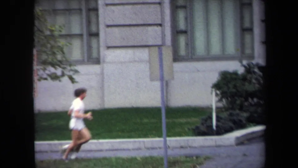 1970 Washington, DC frame showing a jogger moving past grass, shrubs, a signpost, and a stone building facade.