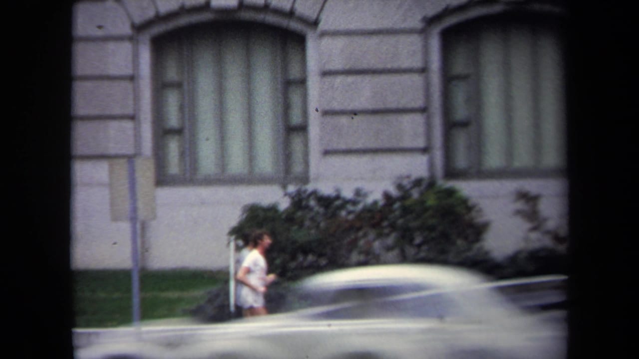 Woman jogging on a sidewalk beside a grey stone building as a car passes in the foreground.