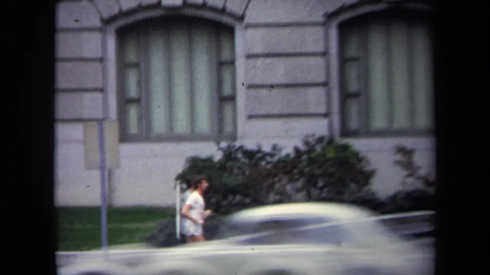 1970 Washington, DC home-movie frame showing a woman jogging past a grey stone building while a light-colored car crosses the foreground.