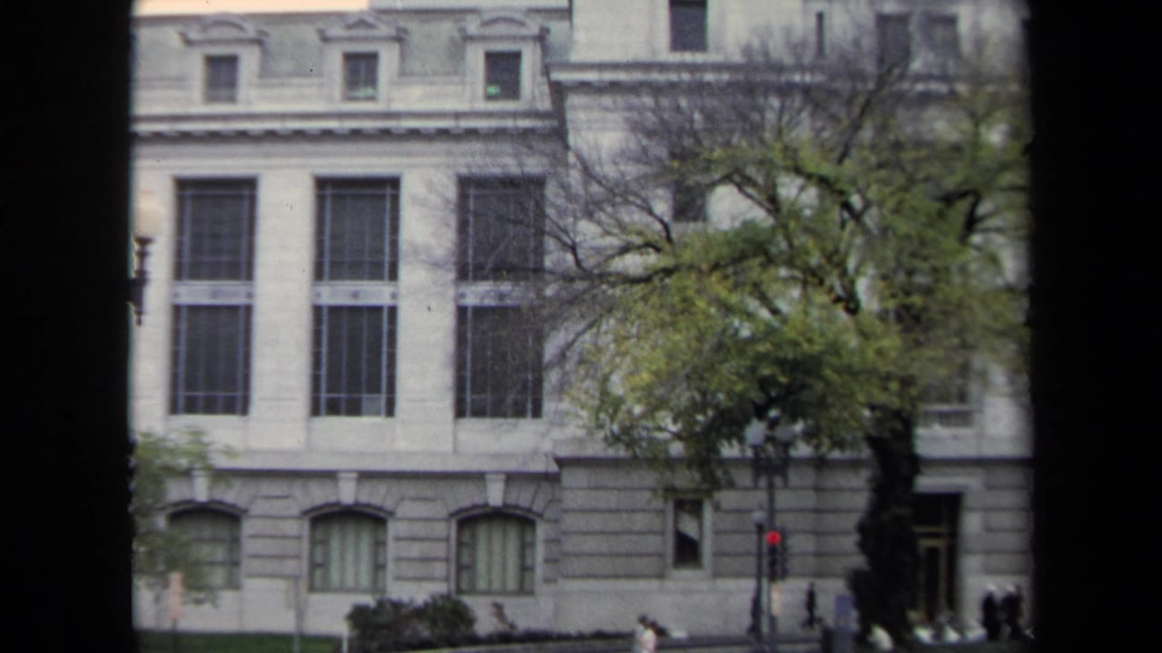 Large grey building with tall windows, a street tree, traffic signal, and pedestrians near the entrance.