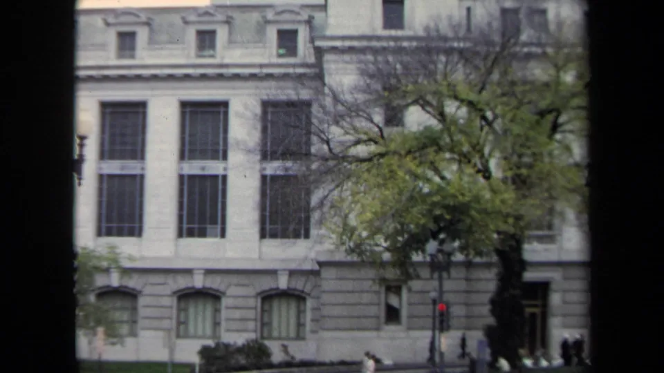 1970 Washington, DC frame of a large grey institutional building with tall windows, a street tree, pedestrians, and a traffic signal.