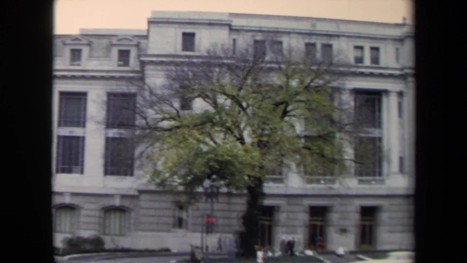 1970 Washington, DC home-movie view of a grey stone building, large tree, traffic signal, pedestrians, and a blurred passing car.