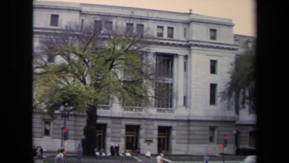 1970 Washington, DC frame showing the grey building facade, large street tree, pedestrians, traffic lights, and the jogger near the bottom edge.