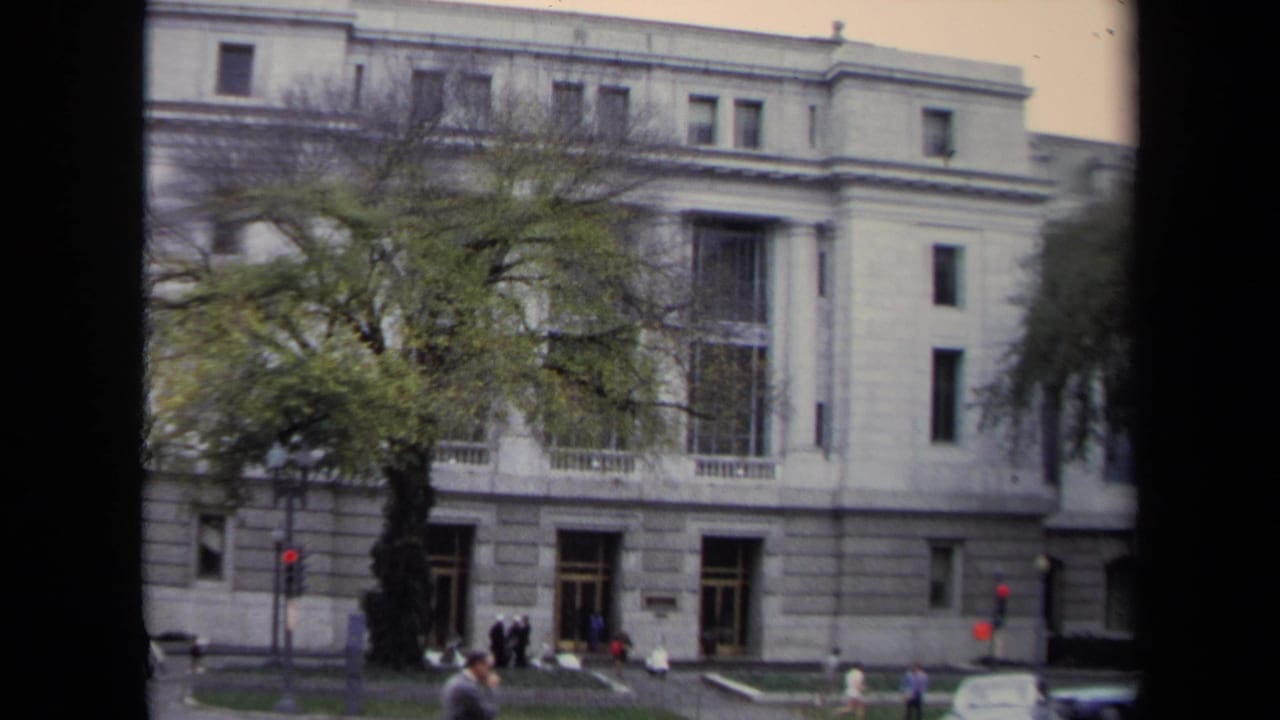 1970 Washington, DC 8mm frame showing a large white columned building, pedestrians, traffic lights, cars, and trees along the street.