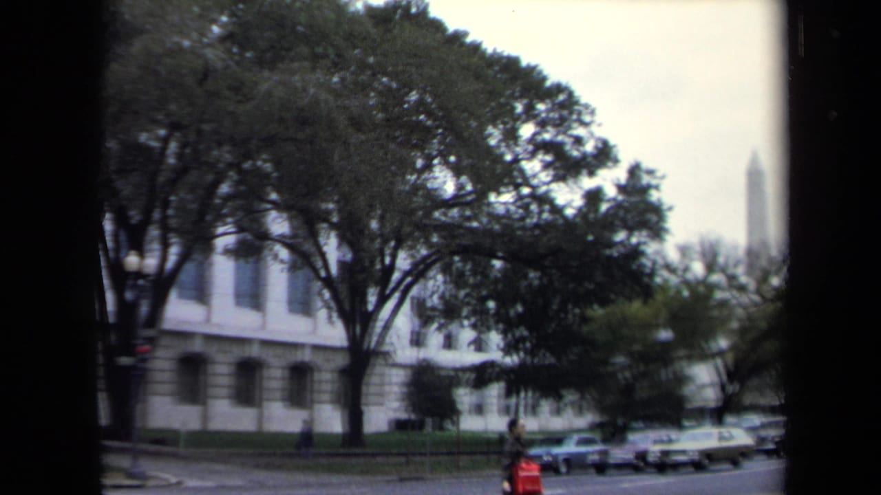 Tree-lined Washington street with cars, a pale building, and the Washington Monument in the distance.