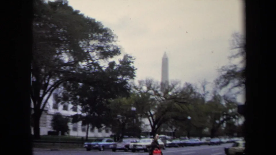 1970 Washington, DC 8mm frame showing traffic and parked cars beneath trees, with pale buildings and the Washington Monument beyond.