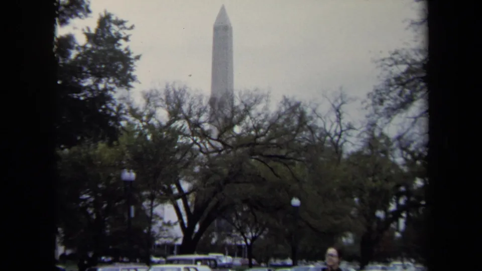1970 Washington, DC view of the Washington Monument above bare and leafy trees, with cars and a partially visible pedestrian at the street edge.