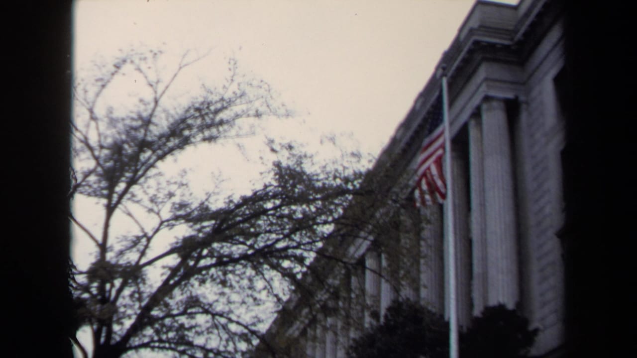 1970 Washington DC home-movie frame showing a classical building, bare trees, shrubs, and an American flag at half-staff.