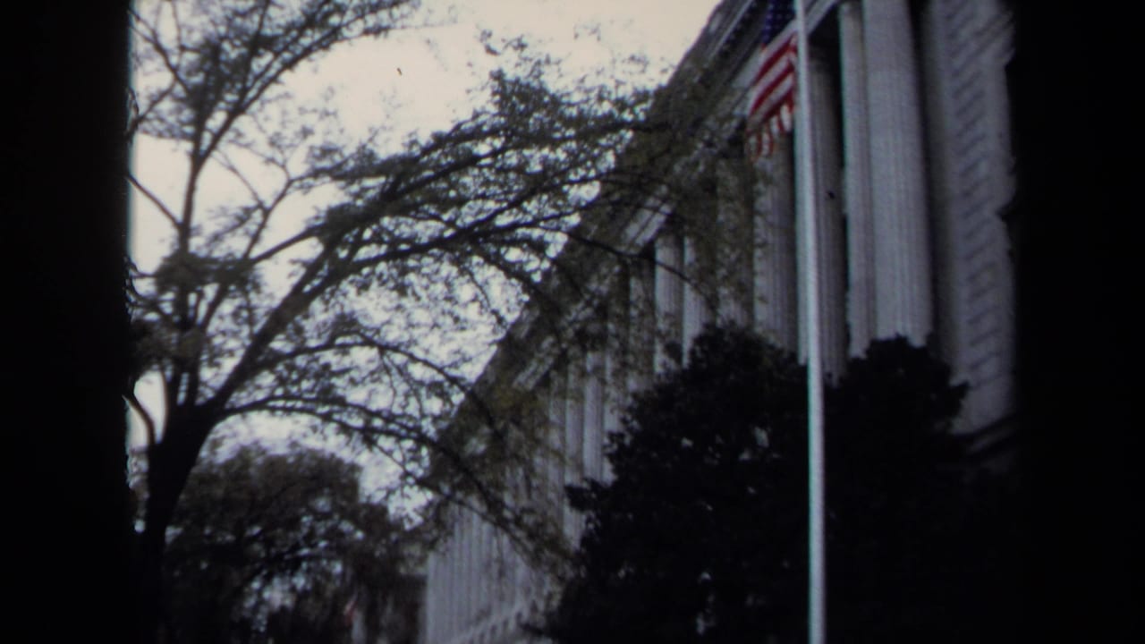 Washington DC exterior view of a formal columned building, trees, shrubs, and a visible American flag on 8mm film.