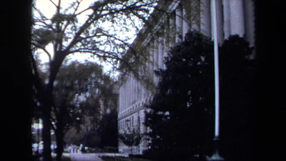 1970 Washington DC street-level view along a sidewalk beside a formal building, with trees, shrubs, and distant pedestrians or cars.