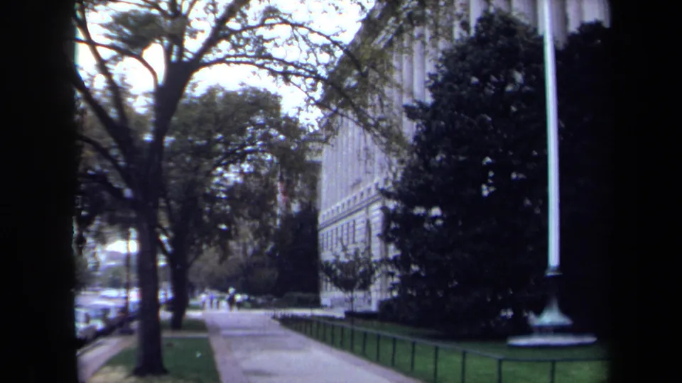 Washington DC home-movie frame showing a sidewalk beside a classical building, trees, parked cars, lawn, and a flagpole.