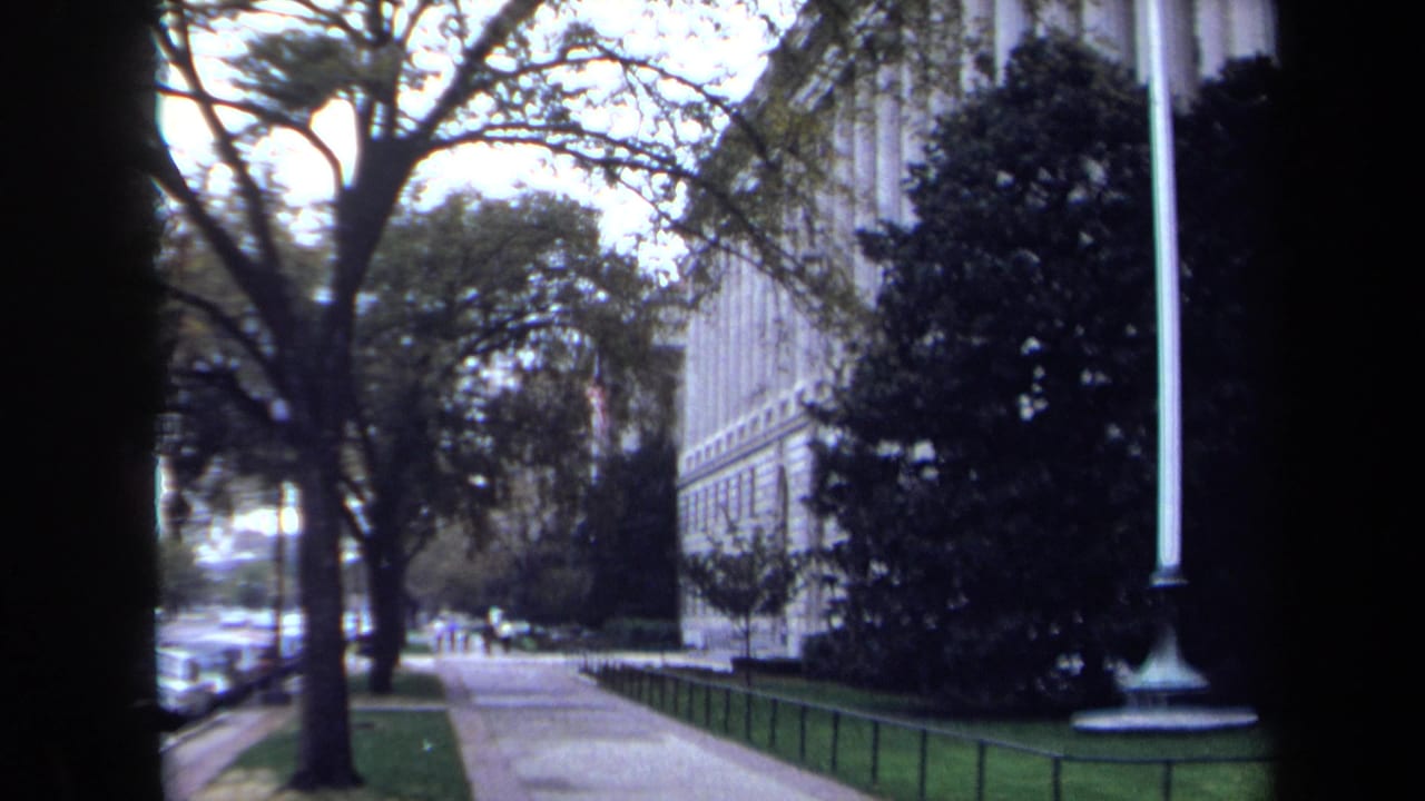 Sidewalk view beside a large stone building with trees and a tall flagpole.