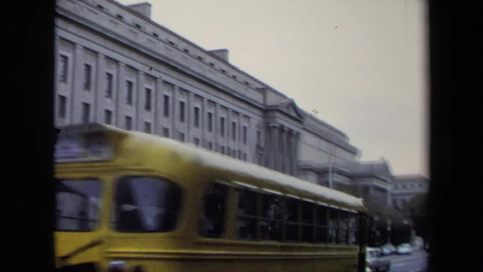 A yellow school bus moves past a large Washington, DC government-style building in this 1970 8mm school-trip view.