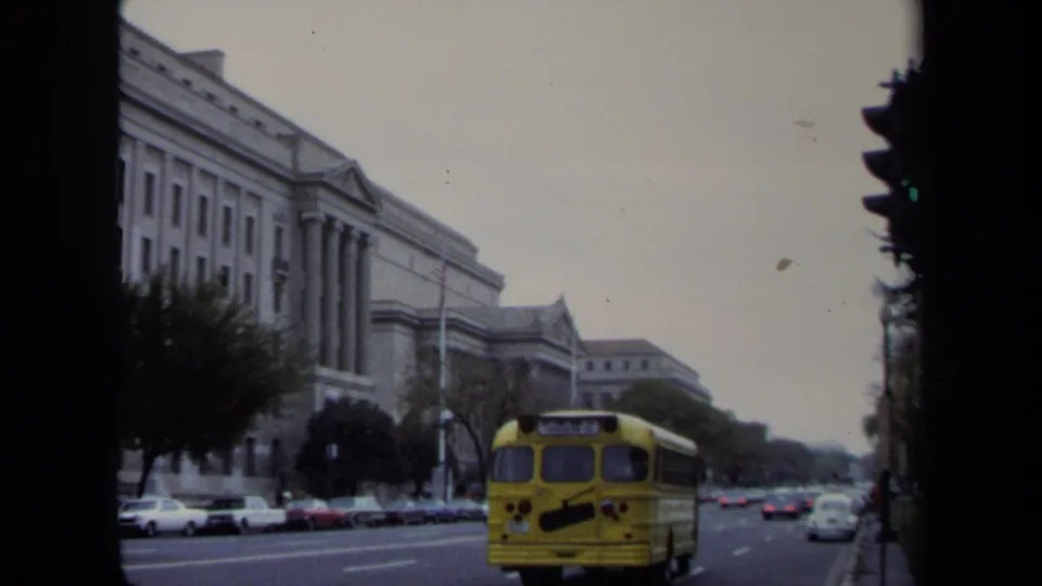 Rear view of a yellow school bus driving along a Washington, DC street with a columned stone building, parked cars, and an American flag visible.