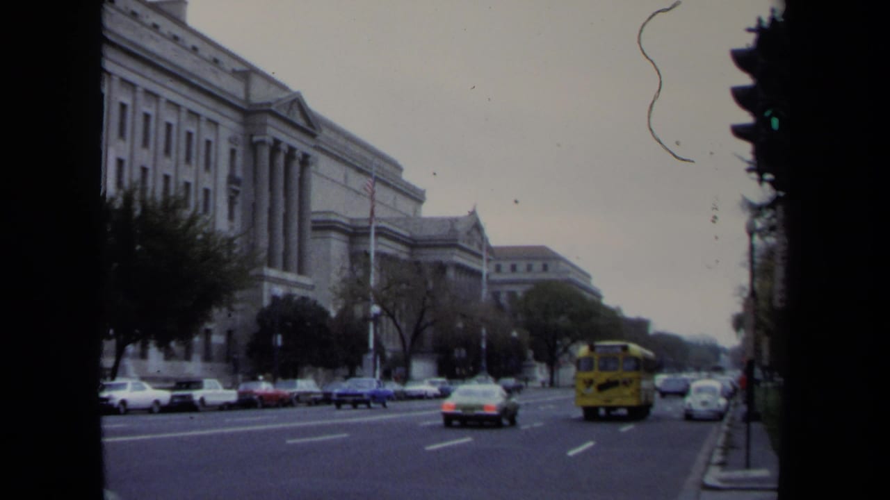 Traffic moves along a city avenue with a yellow school bus near large public buildings.
