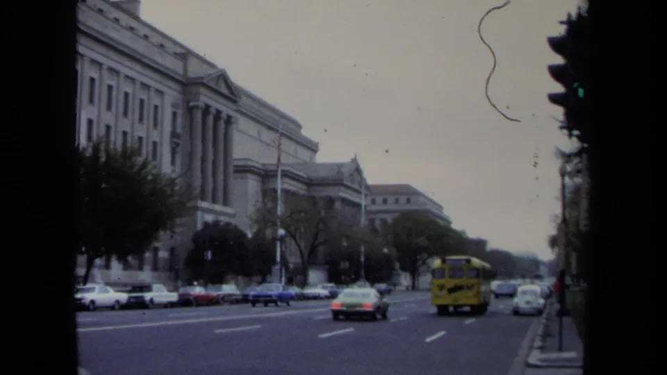 Washington, DC avenue traffic in 1970, with classic cars, a yellow school bus, trees, and large civic buildings in view.