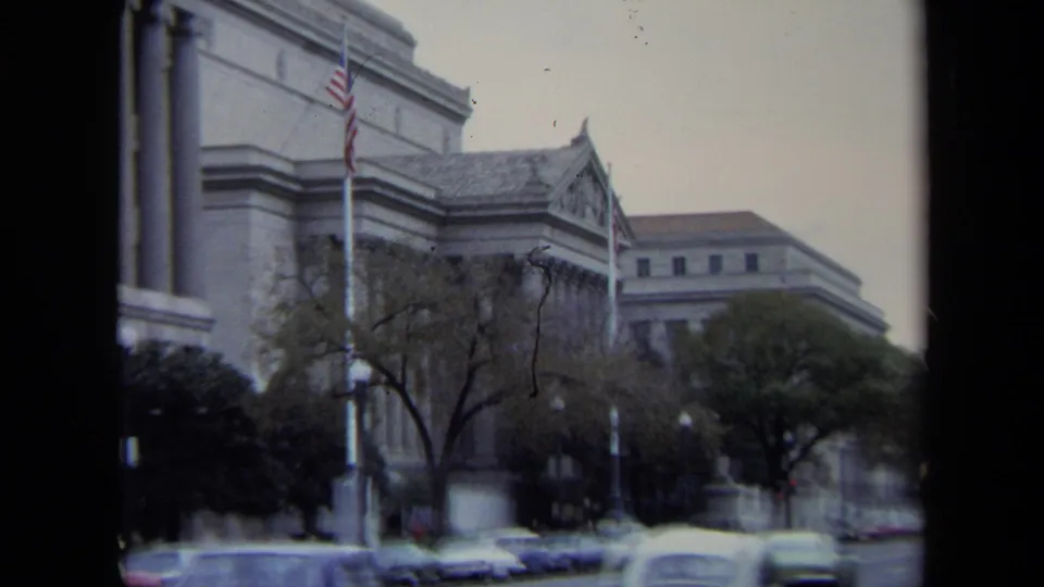 Closer 1970 Washington, DC view of a monumental stone building, flagpole, street trees, and parked classic cars.