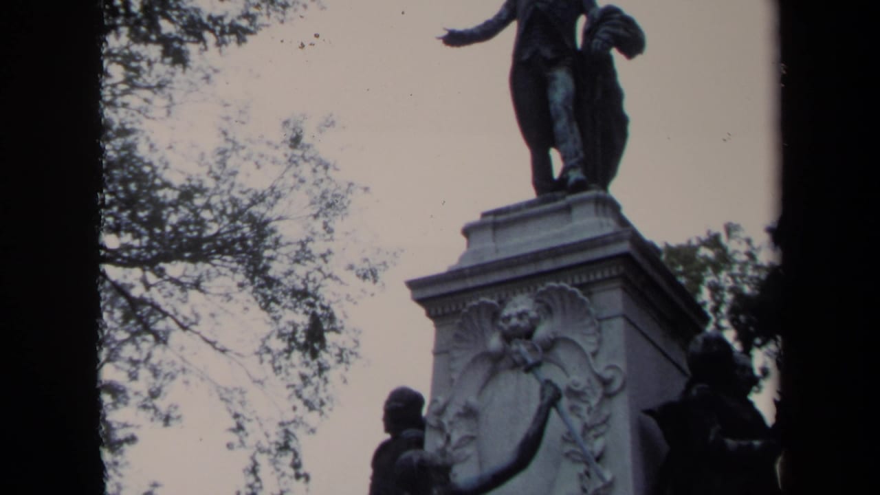 1970 Washington DC 8mm frame showing a bronze statue on a carved stone pedestal, partly obscured by trees and film edge shadows.