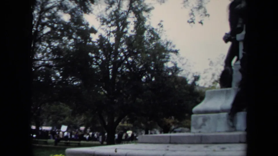 Handheld 1970 Washington DC 8mm view of the monument base with trees, grass, walkways, and a distant gathering on the lawn.