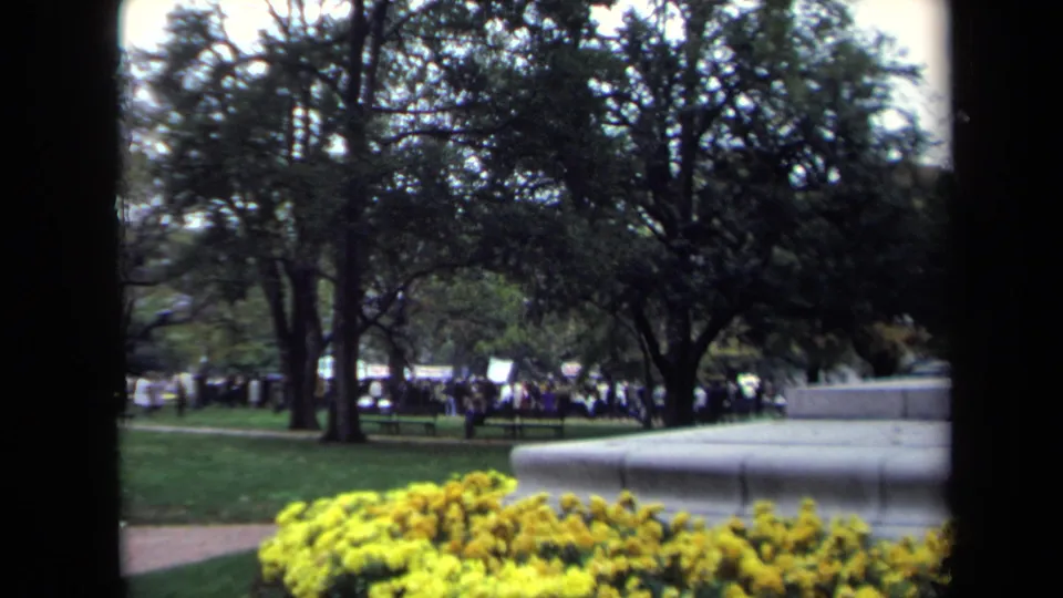 1970 Washington DC 8mm frame with yellow flowers in front of a stone monument base, trees, benches, and a distant gathered crowd.