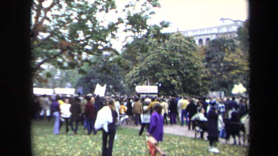 Washington, DC protest crowd in 1970 seen from a handheld 8mm viewpoint, with a banner, trees, and a civic building beyond.