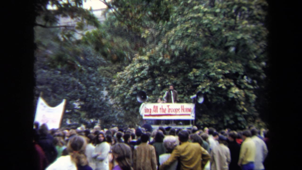 1970 Washington, DC crowd watches a speaker on a platform with a Bring All the Troops Home banner in a leafy outdoor setting.