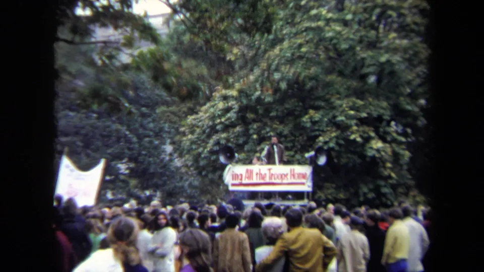 1970 Washington, DC crowd watches a speaker on a platform with a Bring All the Troops Home banner in a leafy outdoor setting.