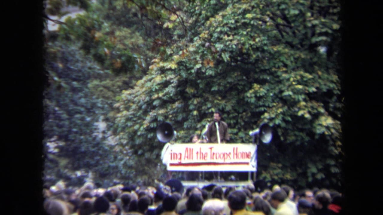 Speaker on a small platform above a dense outdoor crowd with large horn speakers.