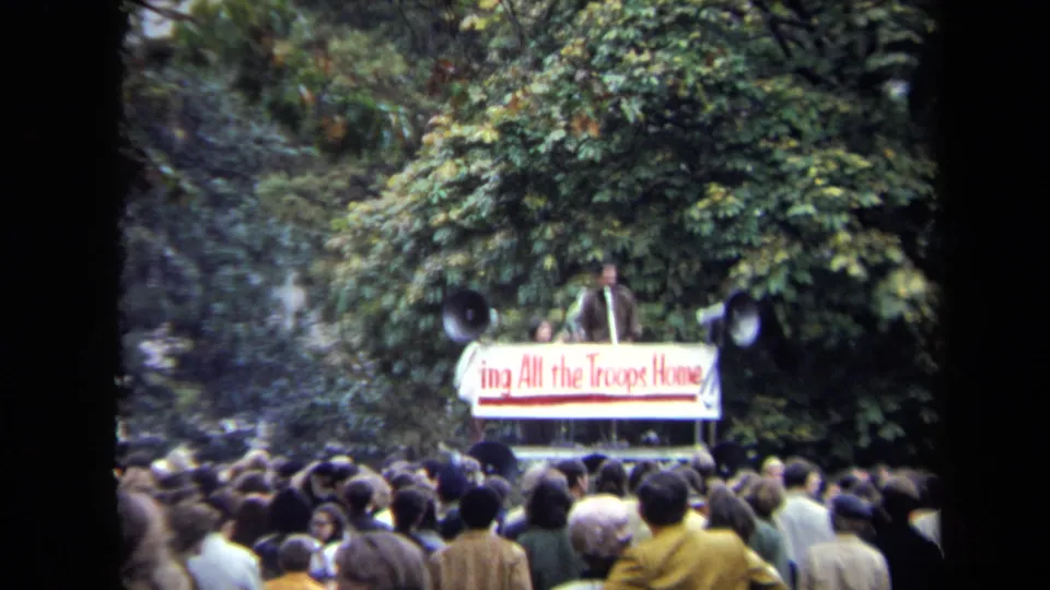 The handheld 8mm frame shows people gathered in Washington, DC, listening to a speaker near a Bring All the Troops Home banner.