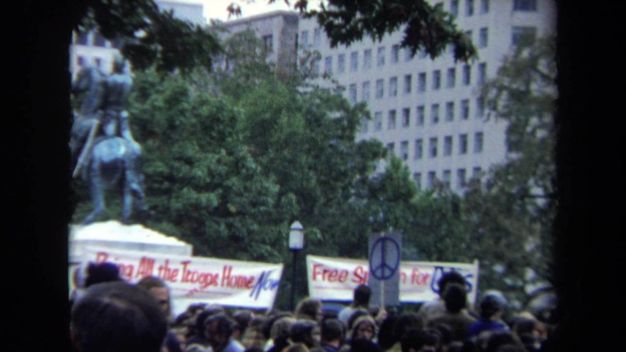 1970 Washington, DC protest crowd viewed from behind, with banners, a peace symbol sign, trees, a statue, and office buildings visible.