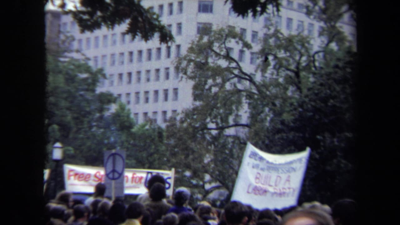 Protest crowd with a peace sign and a white banner beside trees and buildings.