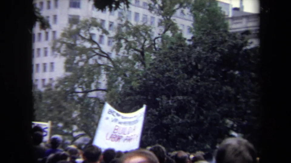 1970 Washington, DC home-movie frame showing a crowd beneath a white protest banner, with trees and a large building in the background.