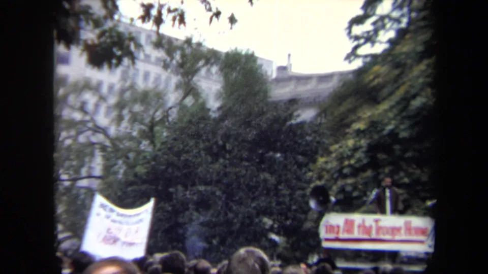 Washington, DC protest scene with a crowd, white banner, trees, buildings, loudspeakers, and a sign on a platform or vehicle.
