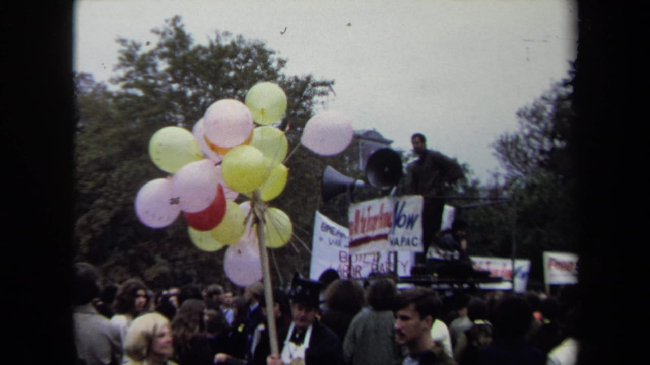 Crowd at a protest with pastel balloons, signs, and a speaker on a raised platform.