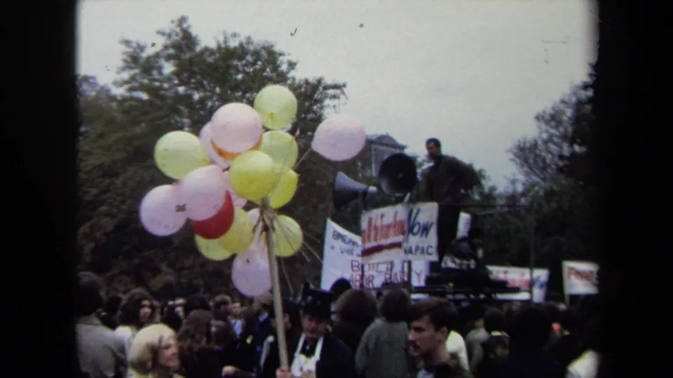 1970 Washington, DC protest crowd with pastel balloons in the foreground and a speaker using a megaphone from a raised platform.